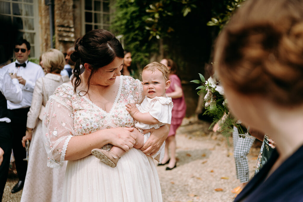 Mother in a floral lace dress holding a baby outdoors at a wedding, smiling at the child. Andrew Brannan Photography