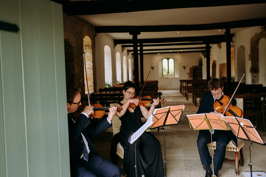 Three violinists in formal attire perform chamber music in a stone-walled chapel with arched windows to the back. Andrew Brannan Photography