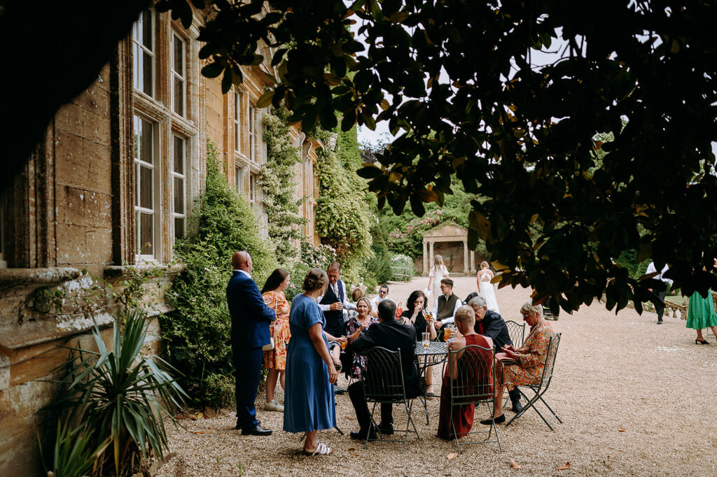 Guests at an outdoor wedding reception gather around a metal table in a gravel courtyard beside a historic stone building with leafy trees overhead. Andrew Brannan Photography