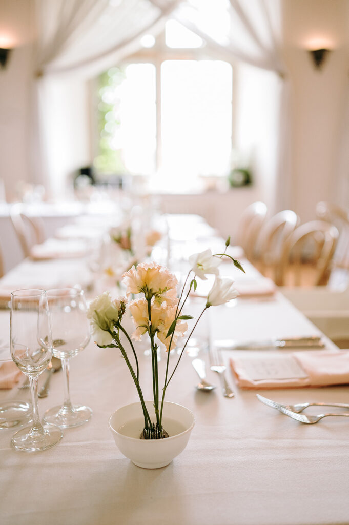 Close-up of a white vase with pale pink flowers on a banquet table, with wine glasses and silverware in a softly lit dining room. Andrew Brannan Photography