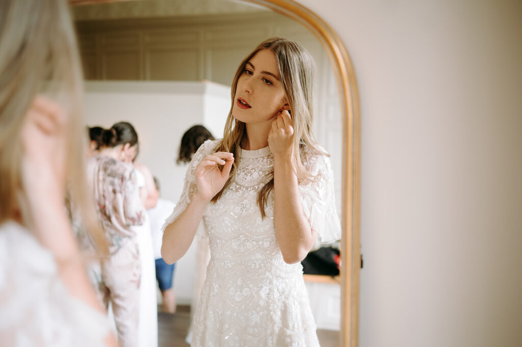 Bride in a lace wedding dress trying on earrings in front of a mirror in a bridal fitting room. Andrew Brannan Photography