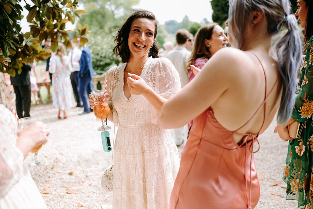 Smiling woman in a white lace dress holds a pink cocktail at an outdoor wedding reception, chatting with friends. Andrew Brannan Photography
