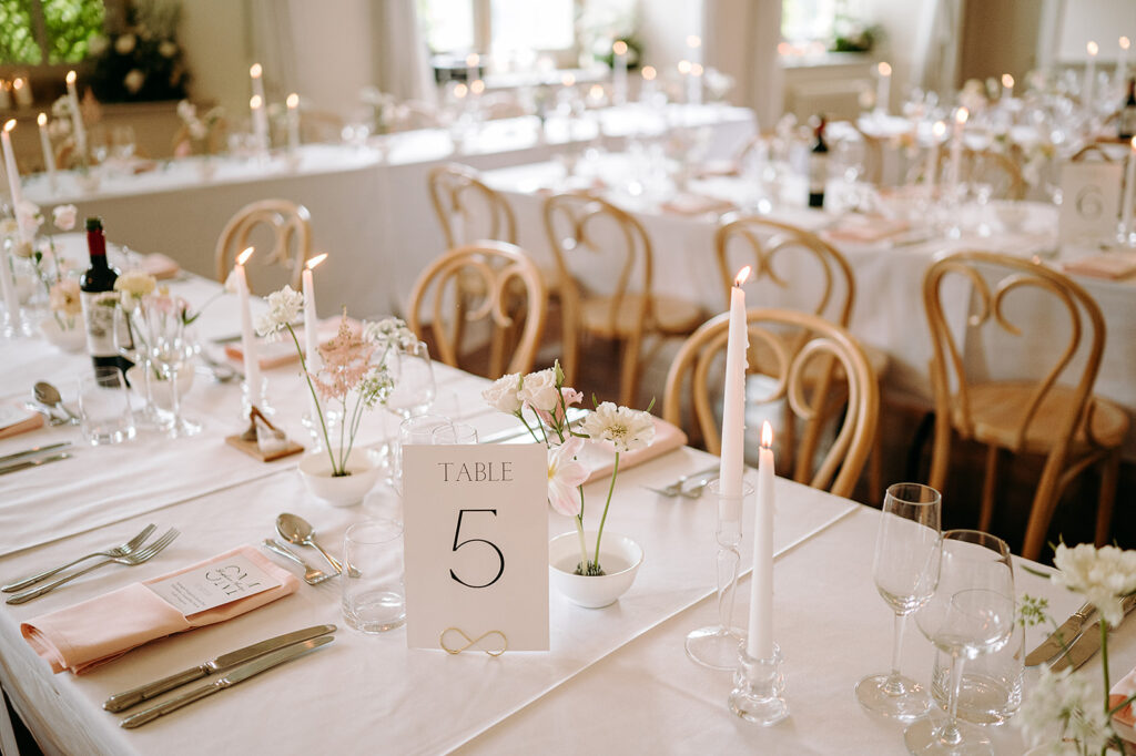 Elegant banquet table set with candles, soft pink flowers, glassware, and a TABLE 5 place card on white linens Andrew Brannan Photography