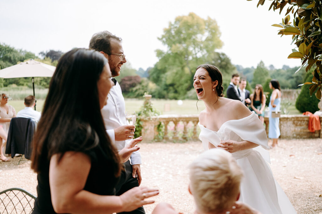 Bride in an off-shoulder white dress laughing with guests at an outdoor wedding reception, holding a glass of champagne. Andrew Brannan Photography