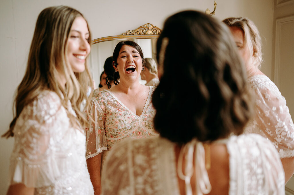 Woman in floral lace dress laughing with bridesmaids in a room with an ornate mirror behind her on the wall. Andrew Brannan Photography