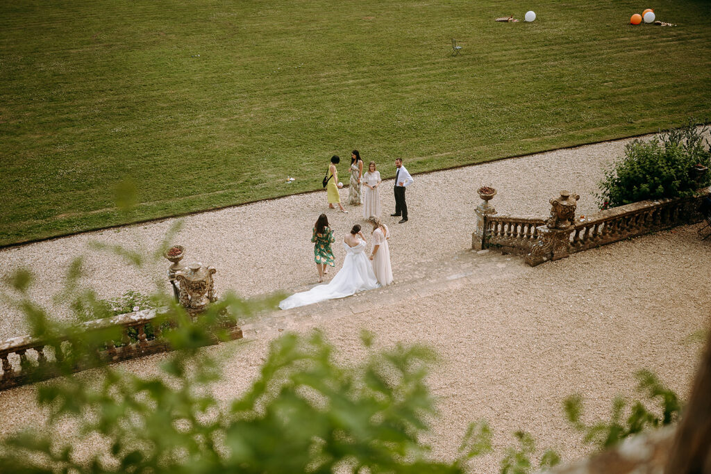 Wedding party on a gravel terrace beside a green lawn; bride in a white gown with a long train, bridesmaids in pastel dresses, guests chatting nearby. Andrew Brannan Photography