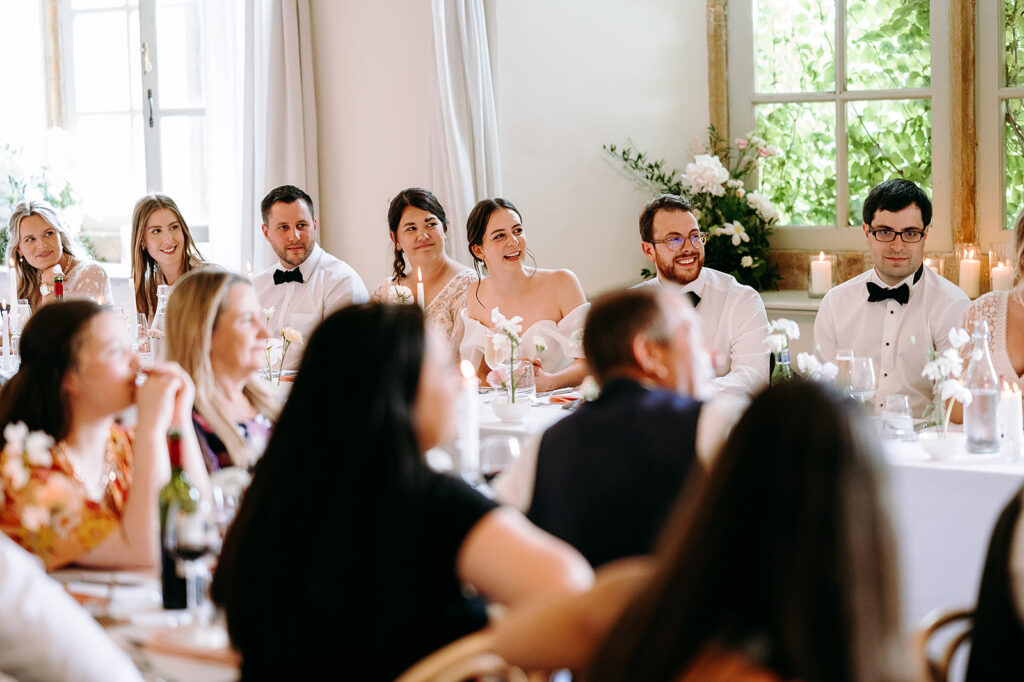 Group of wedding guests in formal attire seated at a long table with candles and white floral centerpieces in a bright room by large windows. Andrew Brannan Photography