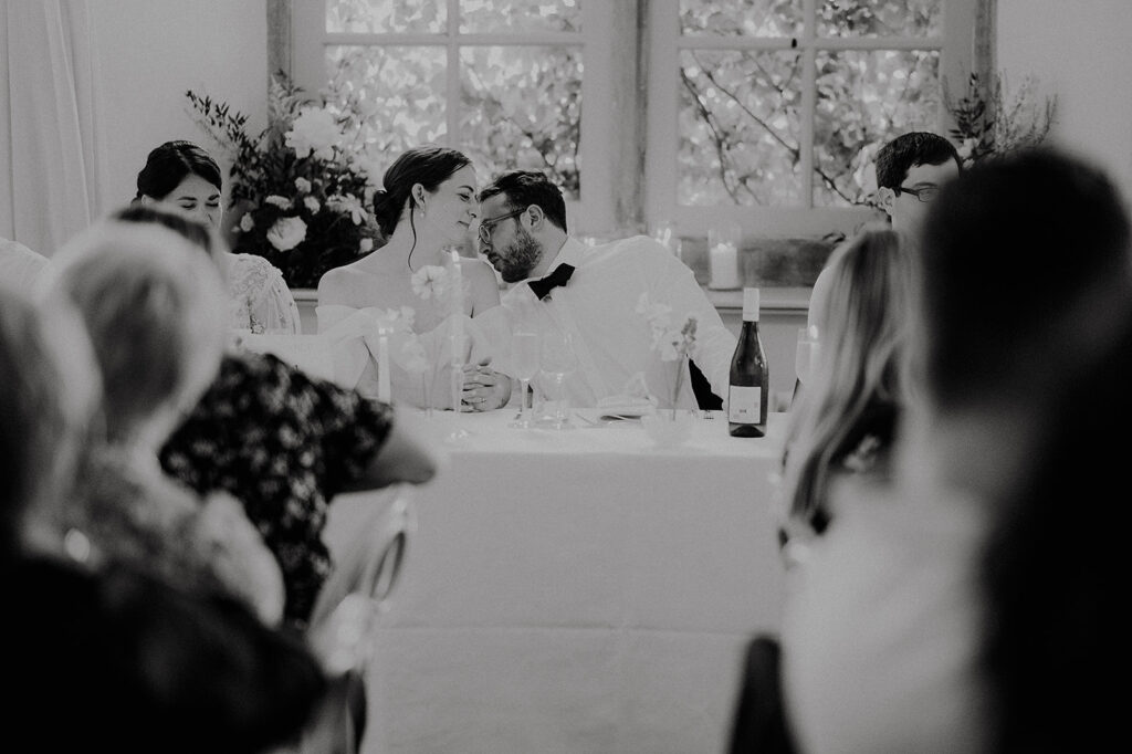 Bride and groom share an intimate moment at their reception, leaning toward each other across a table with wine and flowers nearby. Andrew Brannan Photography