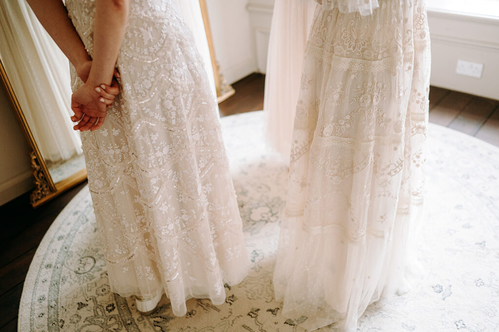 Two brides in ivory lace wedding dresses stand with their hands clasped behind their backs in a bright room with a patterned rug. Andrew Brannan Photography