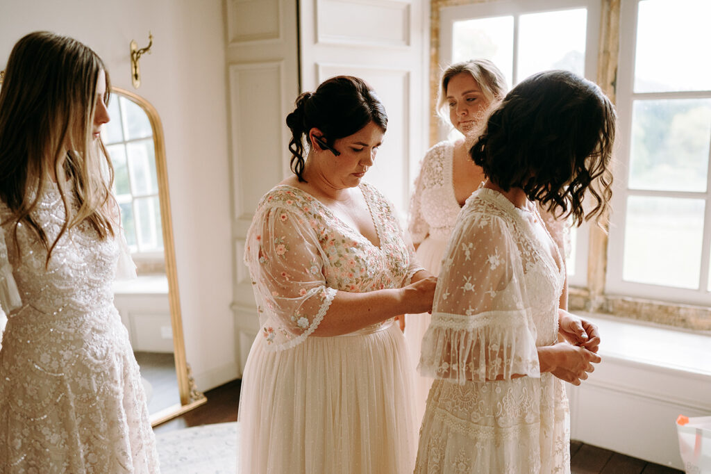 Four bridesmaids in delicate white lace gowns in a sunlit room; one helps another fasten her bodice to prepare for the wedding. Andrew Brannan Photography