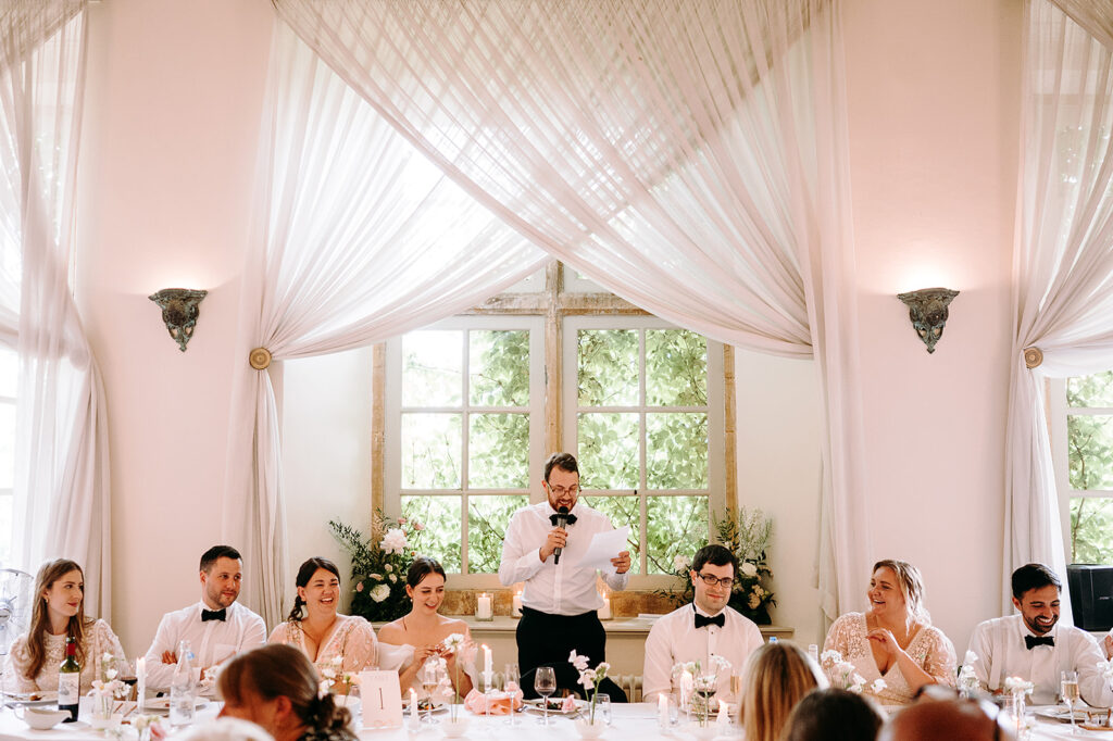Man giving a wedding toast at the head table, reading from a paper as guests listen in a sunlit room with pink drapery and floral decor behind him. Andrew Brannan Photography