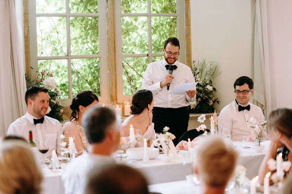 Man in a white shirt and black bow tie delivers a speech with a microphone at a candlelit wedding reception. Andrew Brannan Photography