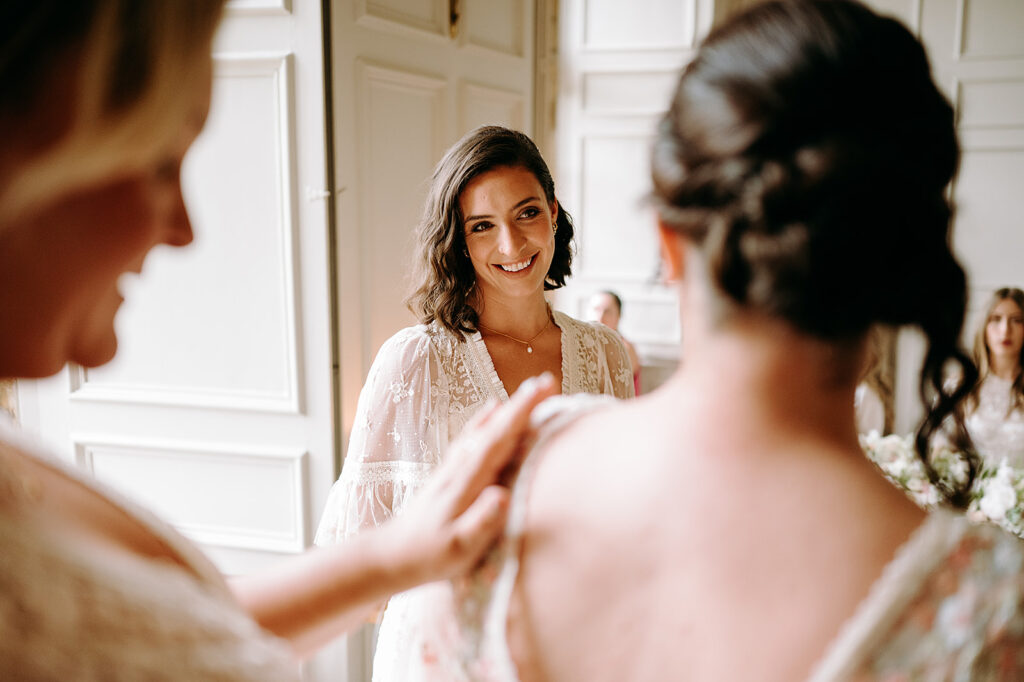 Bride-to-be in a white lace gown smiles at her reflection as friends softly touch her shoulders in a bright, paneled room. Andrew Brannan Photography