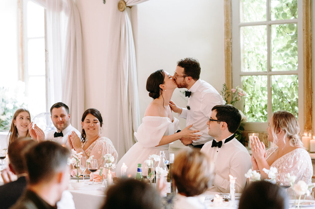 Bride and groom kiss as guests applaud at a wedding reception around a candlelit table. Andrew Brannan Photography