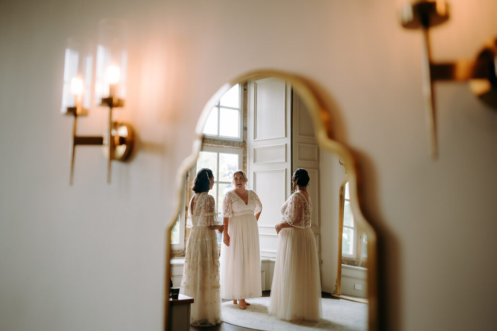 Three women in white wedding dresses stand and chat in a bright, elegant room with large windows, seen through a large arched mirror reflection Andrew Brannan Photography