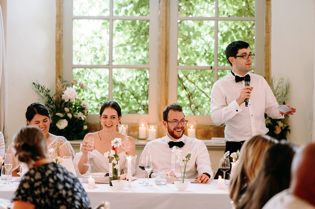 Bride and groom at a decorated wedding reception table, smiling as a man in a white shirt speaks into a microphone nearby. Andrew Brannan Photography