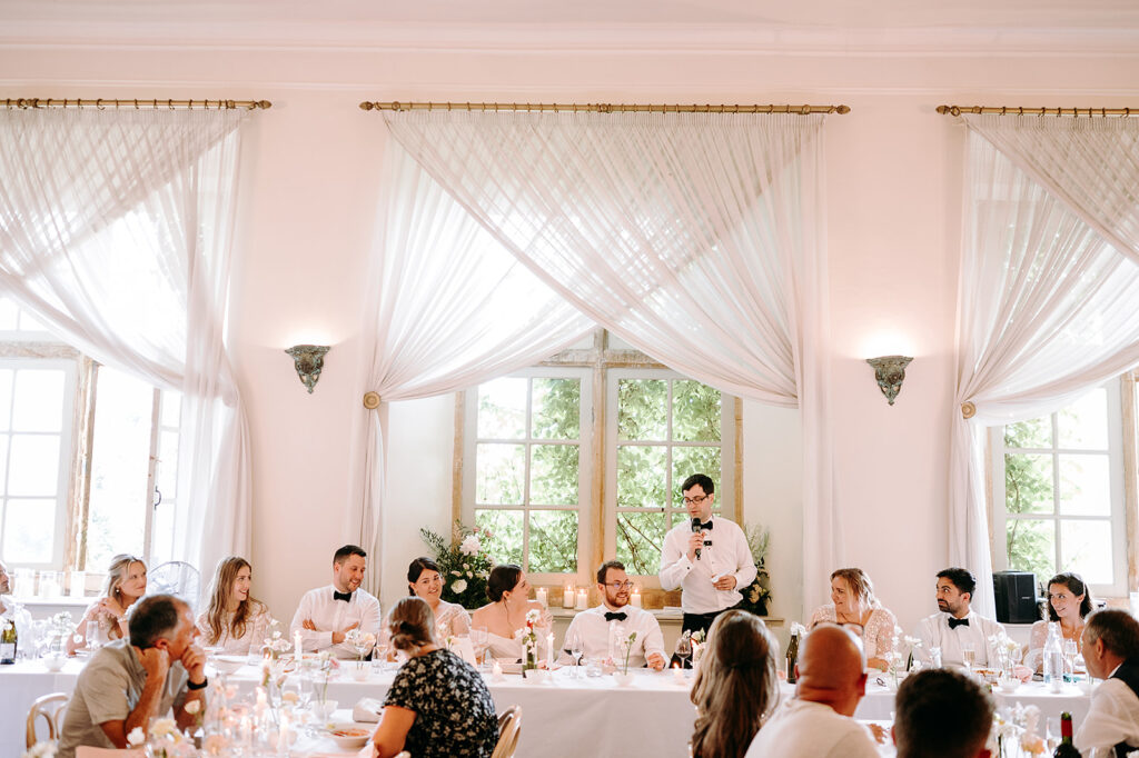 Guests seated at a long reception table as a man in a tuxedo gives a speech with a microphone in a bright, draped room. Andrew Brannan Photography
