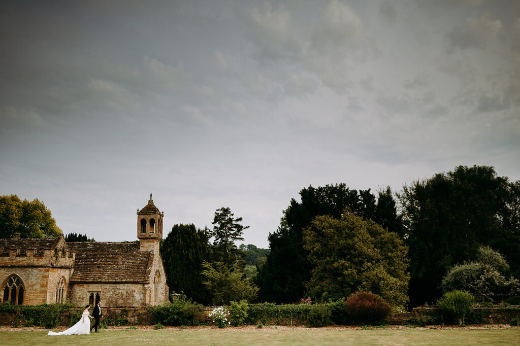 Wedding couple in formal attire walking on a lawn beside a historic stone church with a small bell tower, surrounded by shrubbery and trees under a cloudy sky. Andrew Brannan Photography