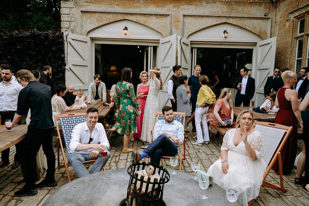 Guests mingle at an outdoor wedding reception beside a rustic stone building with open doors and string lights at dusk/summer. Andrew Brannan Photography