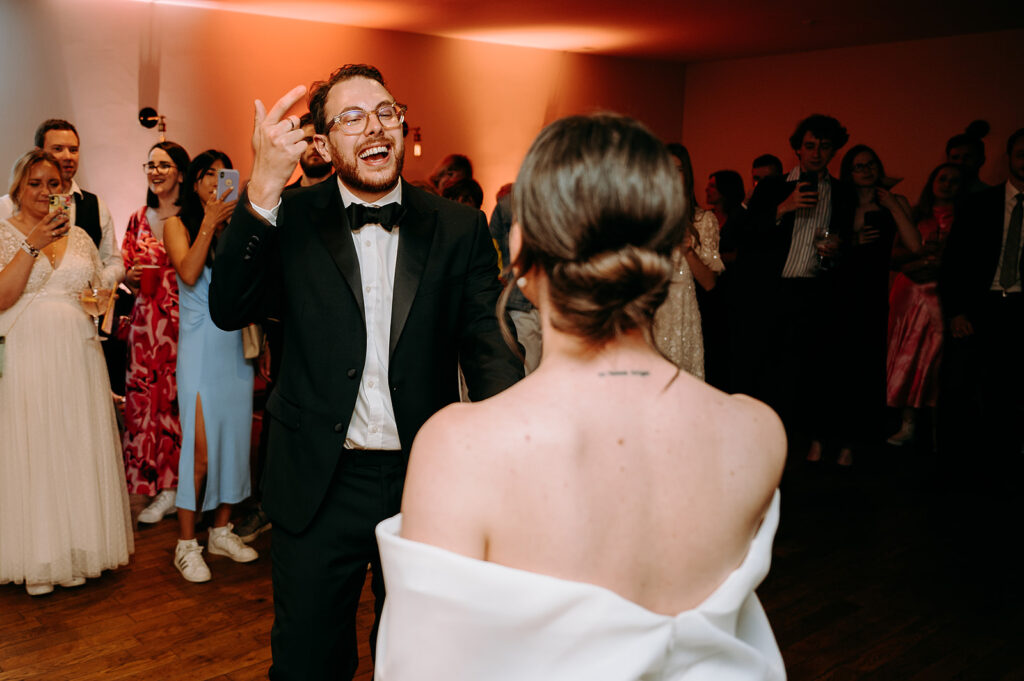 Man in a black tuxedo dancing and laughing toward a woman in a white off-shoulder dress at a wedding reception, guests cheering in the background Andrew Brannan Photography