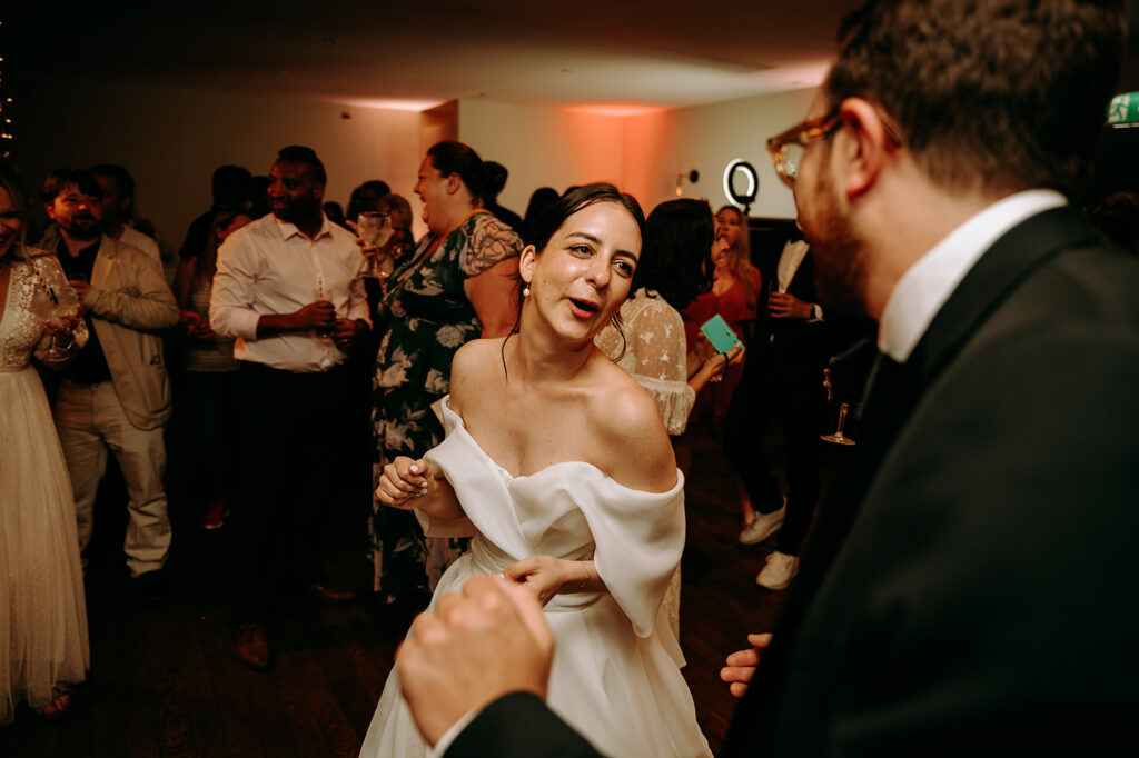 Bride in an off-shoulder white wedding dress dancing with guests at a crowded reception. Andrew Brannan Photography