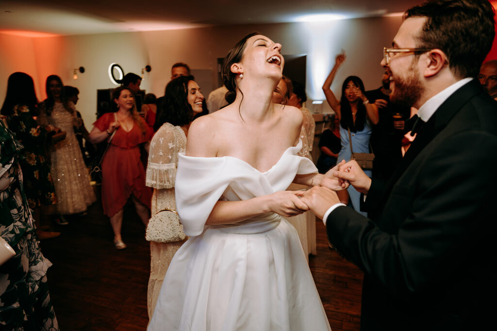 Bride in an off-shoulder white wedding gown laughs while dancing with the groom as guests cheer in the background at a lively reception pass-by scene. Andrew Brannan Photography