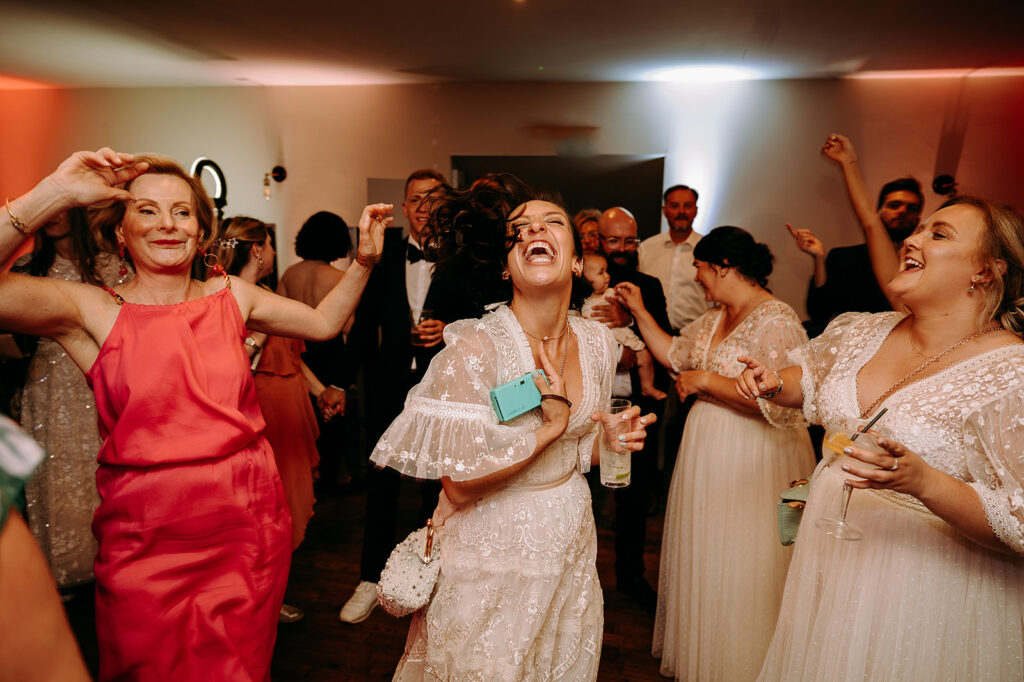 Joyful wedding reception scene with guests dancing; center woman in white lace dress laughing, holding a turquoise phone, surrounded by friends with drinks in a warmly lit room. Andrew Brannan Photography