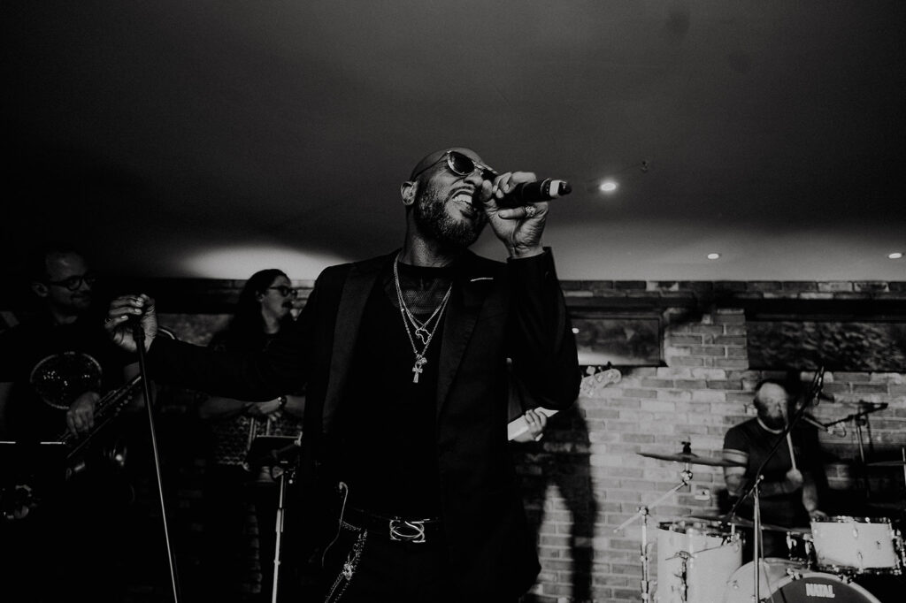 Male singer in a blazer and sunglasses sings into a microphone on stage, band members and drums in the background (black-and-white photo). Andrew Brannan Photography