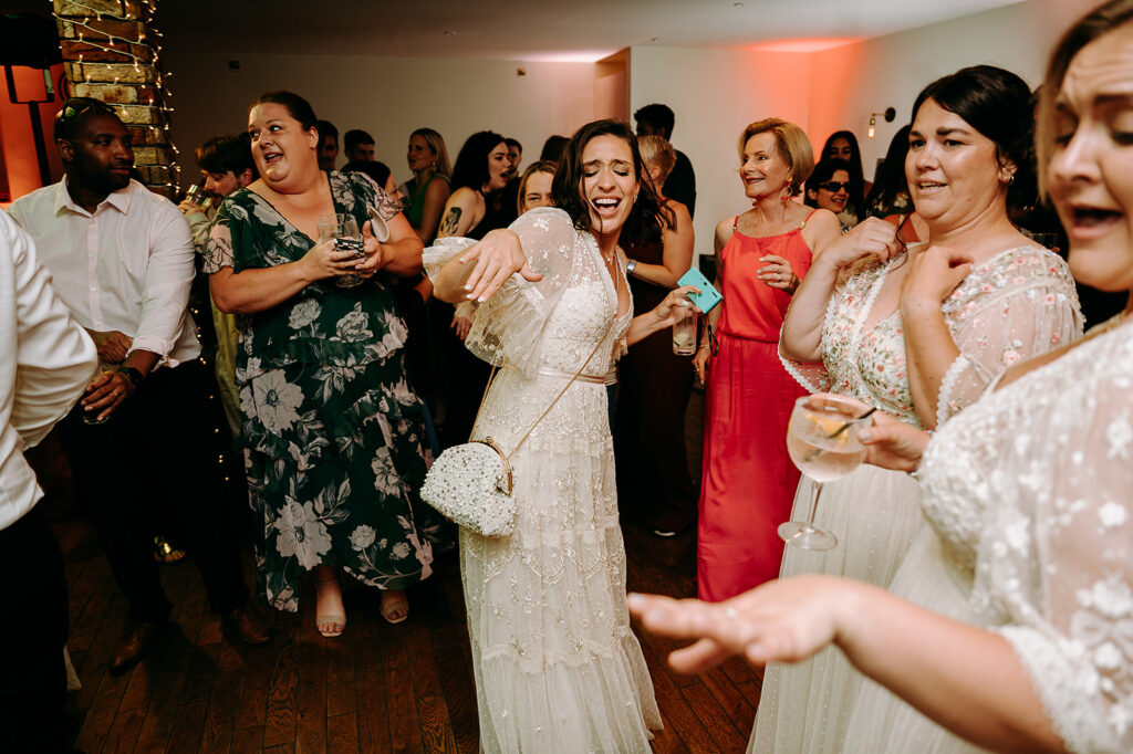 Bride in a white lace dress dancing at a wedding reception, holding a small beaded purse and a drink. Andrew Brannan Photography