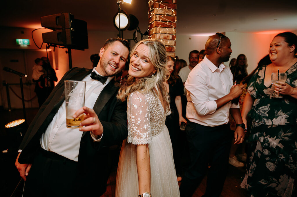 Smiling couple toasting at a wedding reception; man in a tuxedo, woman in a pale lace dress, with guests dancing in the background. Andrew Brannan Photography