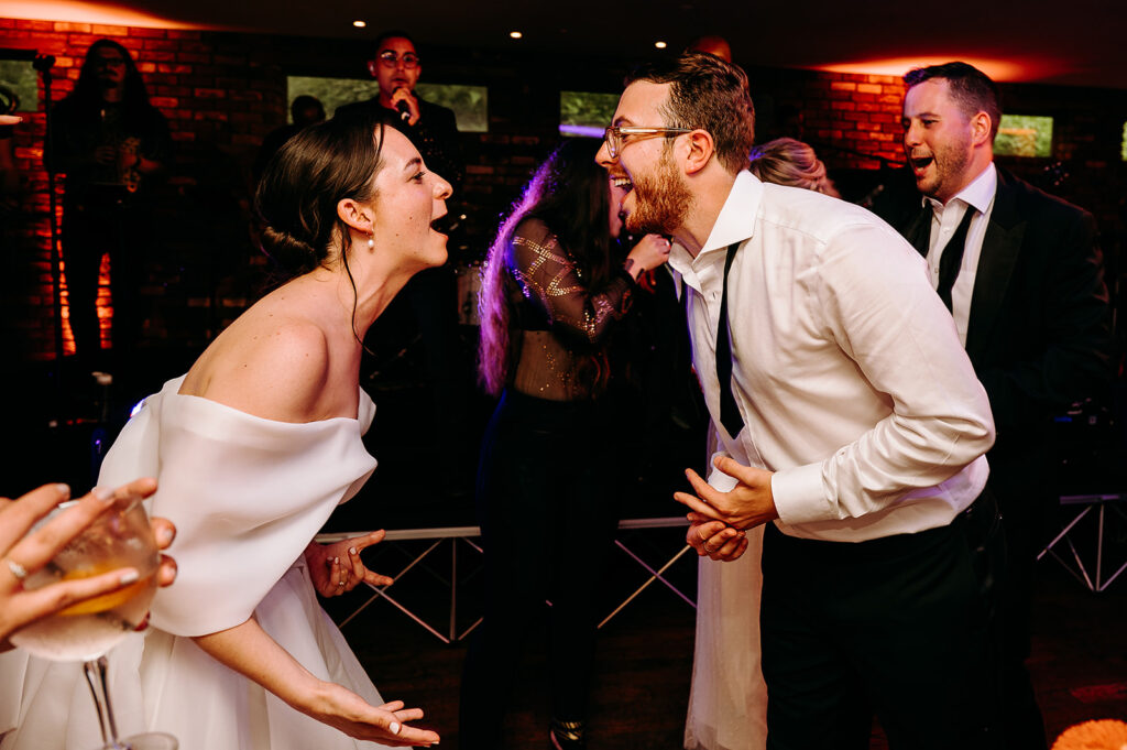Bride in an off‑the‑shoulder white gown and groom in a white shirt laugh and lean toward each other on the dance floor under warm lighting on a wedding night. Andrew Brannan Photography