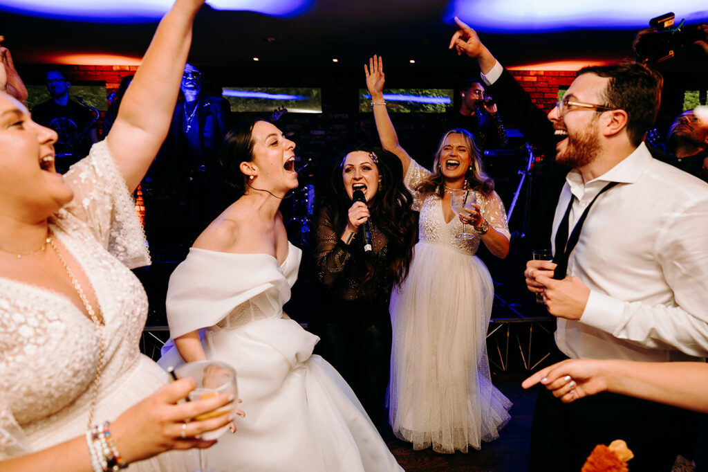 Bride in white gown sings and dances at a lively wedding reception while a woman with a microphone and friends cheer around her. Andrew Brannan Photography