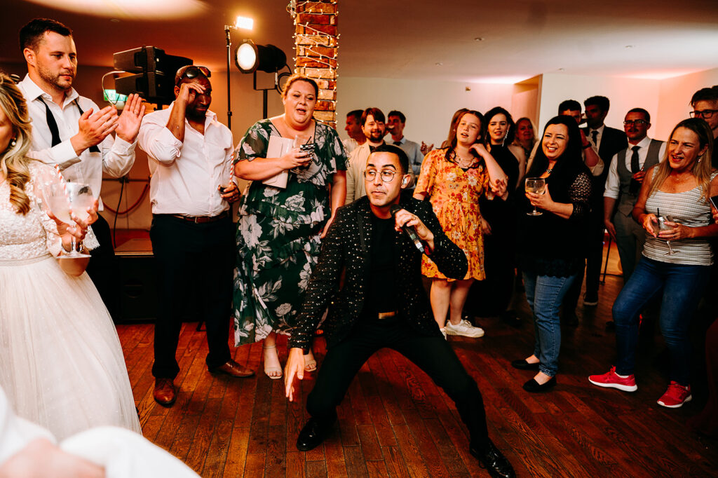Man in a sparkly black jacket singing into a microphone at a party, surrounded by cheering guests in a crowded room. Andrew Brannan Photography
