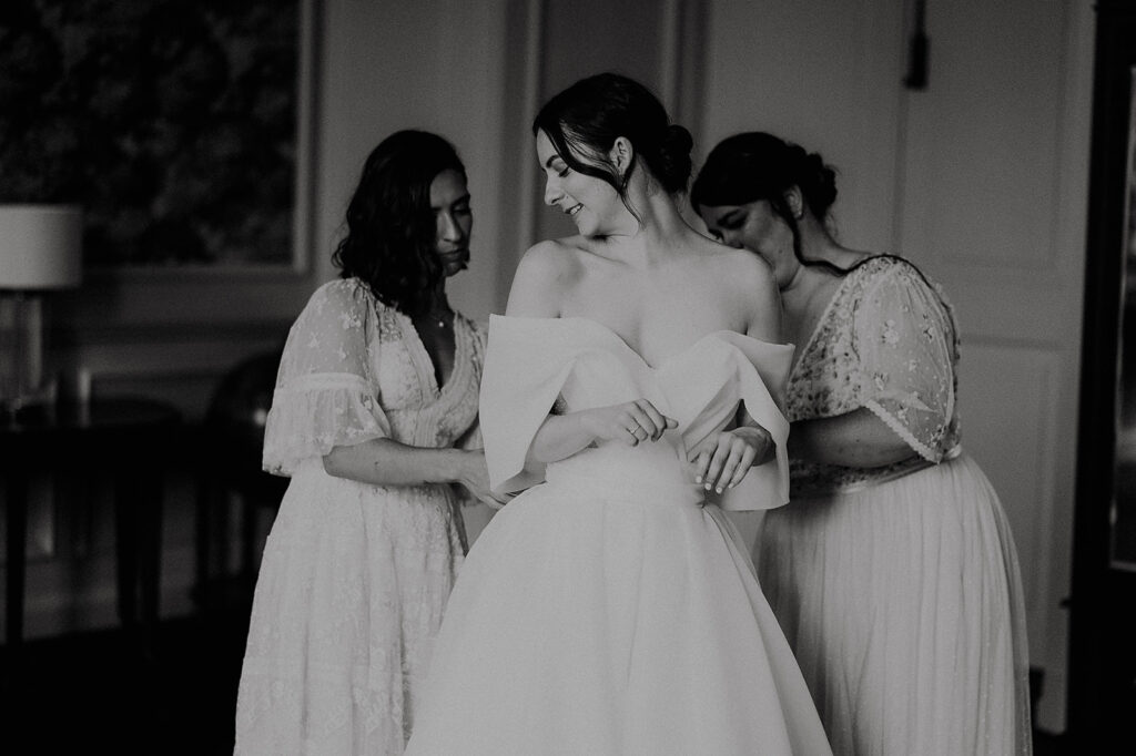 Bride in an off-shoulder gown stands center as two bridesmaids in lace dresses help adjust her dress in a dim room. Andrew Brannan Photography