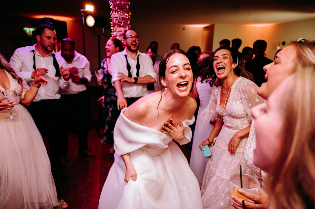 Bride in an off-shoulder white gown laughs and dances at a wedding reception, surrounded by friends in formal attire. Andrew Brannan Photography