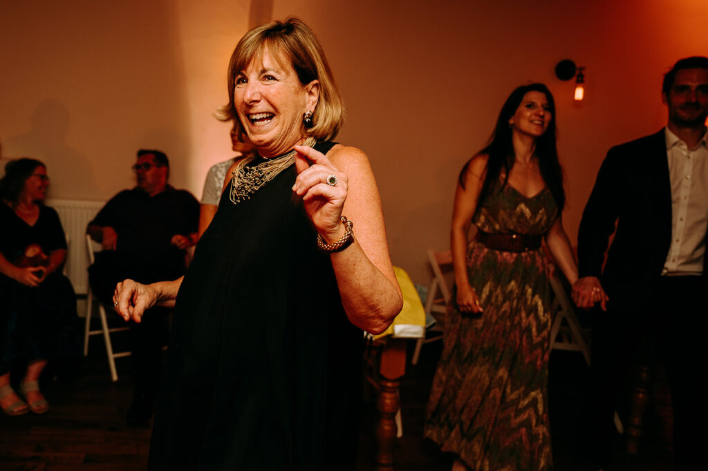 Smiling older woman in a black dress dances at a party, jewelry gleaming, warm orange lighting in the background. Andrew Brannan Photography