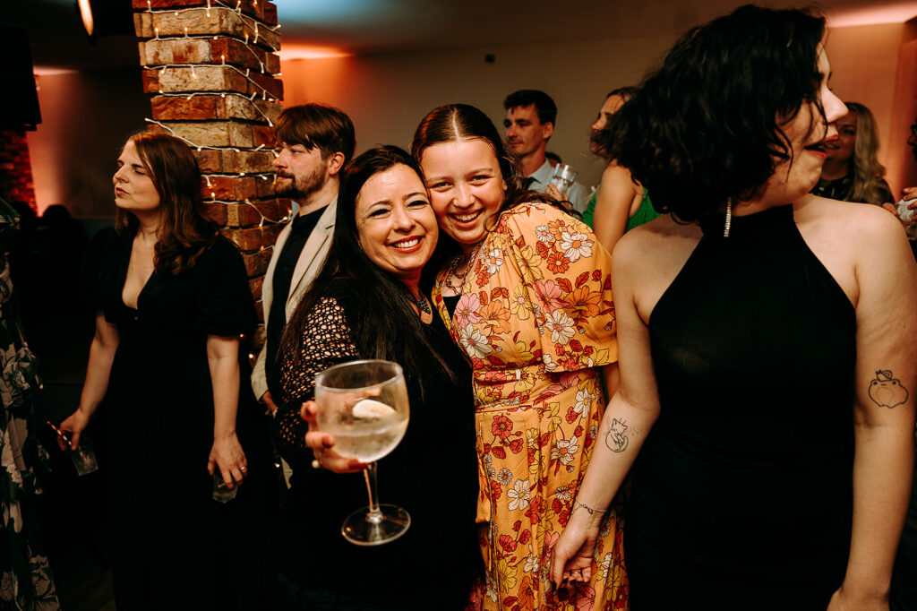 Two smiling women hug at a party, one holding a wine glass, with guests mingling near a brick pillar wrapped in string lights in the background. Andrew Brannan Photography