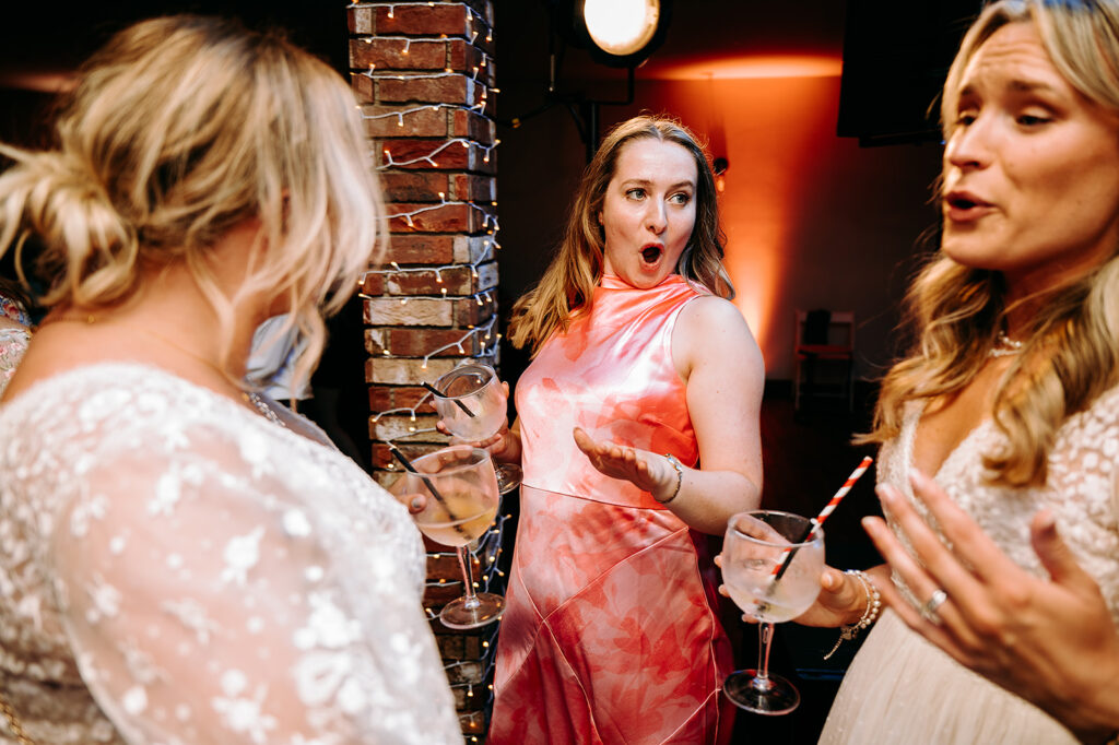 Center woman in pink dress with surprised expression, holding a glass, among friends at a party with string lights overhead. Andrew Brannan Photography