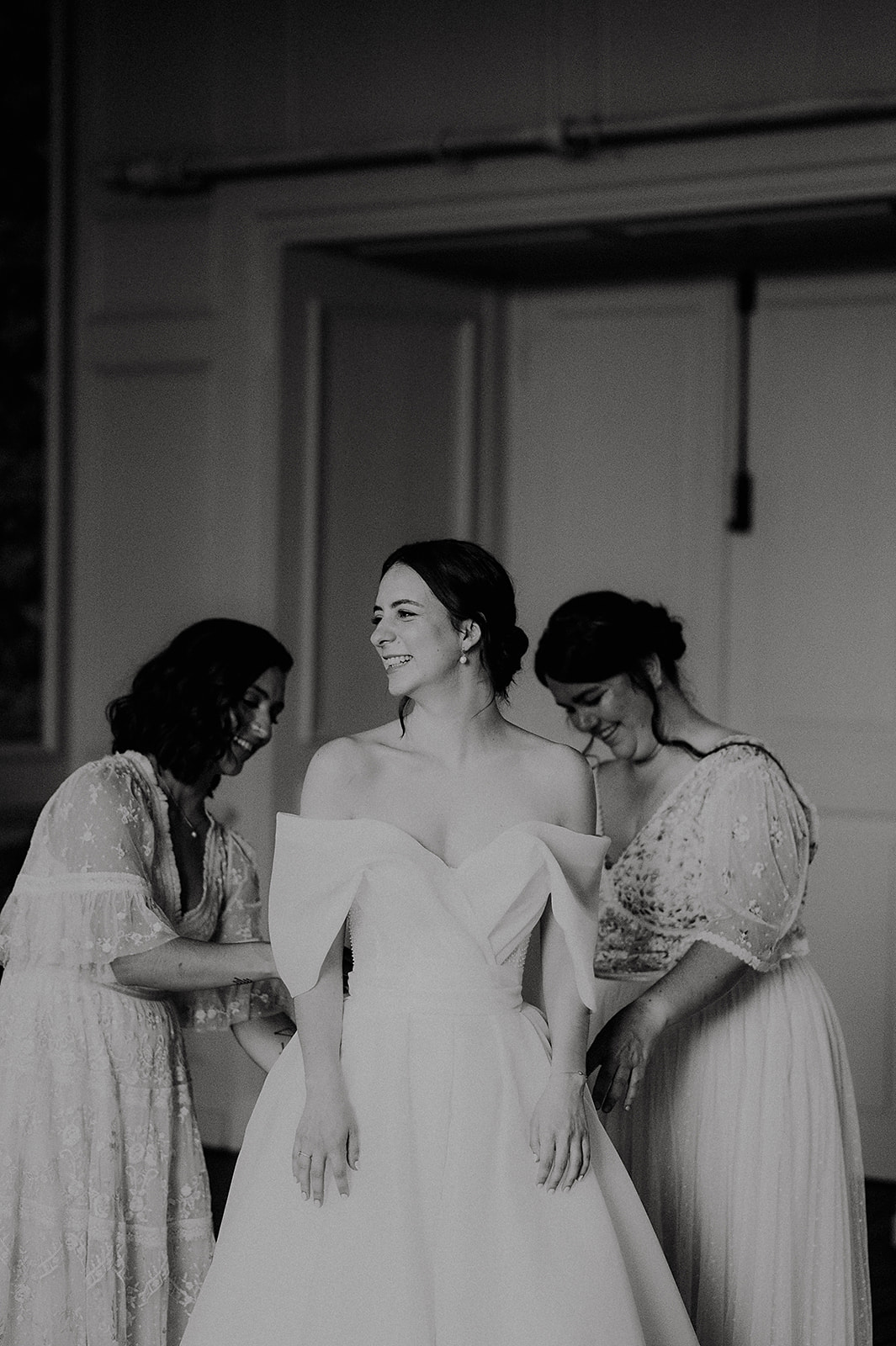 Bride in an off-the-shoulder wedding dress smiles as two bridesmaids adjust her gown. (Black and white) Andrew Brannan Photography