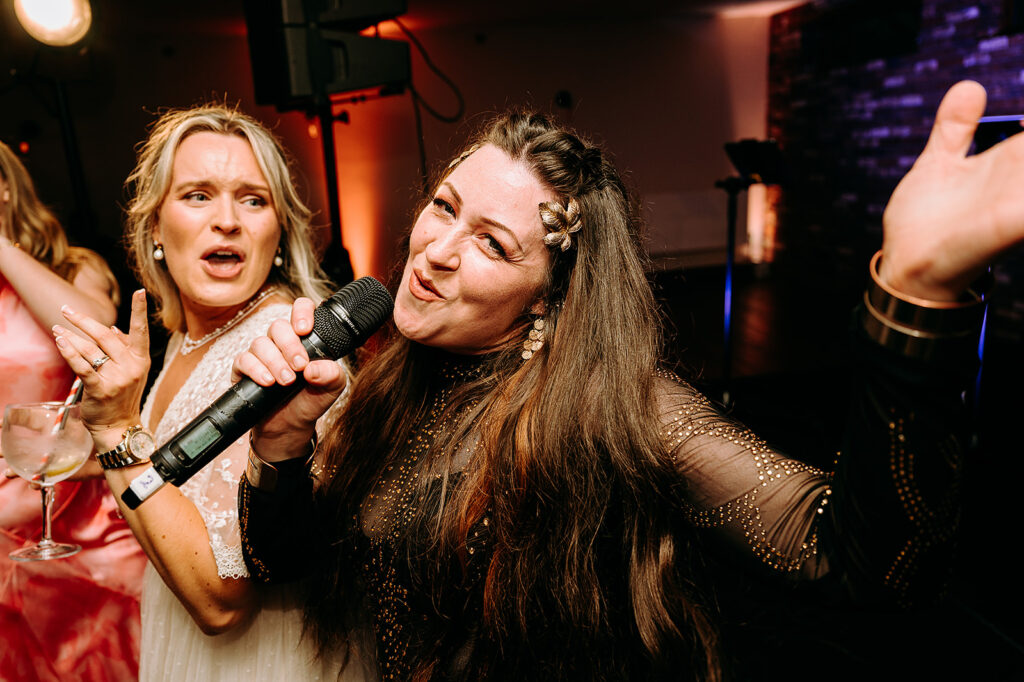 Two women sing into a microphone at a lively party, one in white lace and the other in a glittery black outfit with a gold hair accessory Andrew Brannan Photography