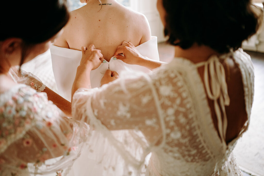 Bride's back as two helpers fasten the back of her off-the-shoulder wedding gown. Andrew Brannan Photography