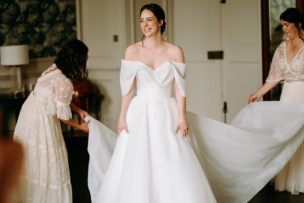 Bride in an off-the-shoulder white wedding gown, smiling as two assistants arrange her train indoors. Andrew Brannan Photography