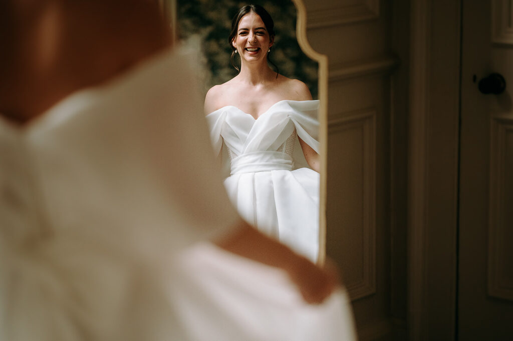 Bride in an off-the-shoulder white wedding gown smiling at her reflection in a tall mirror. Andrew Brannan Photography