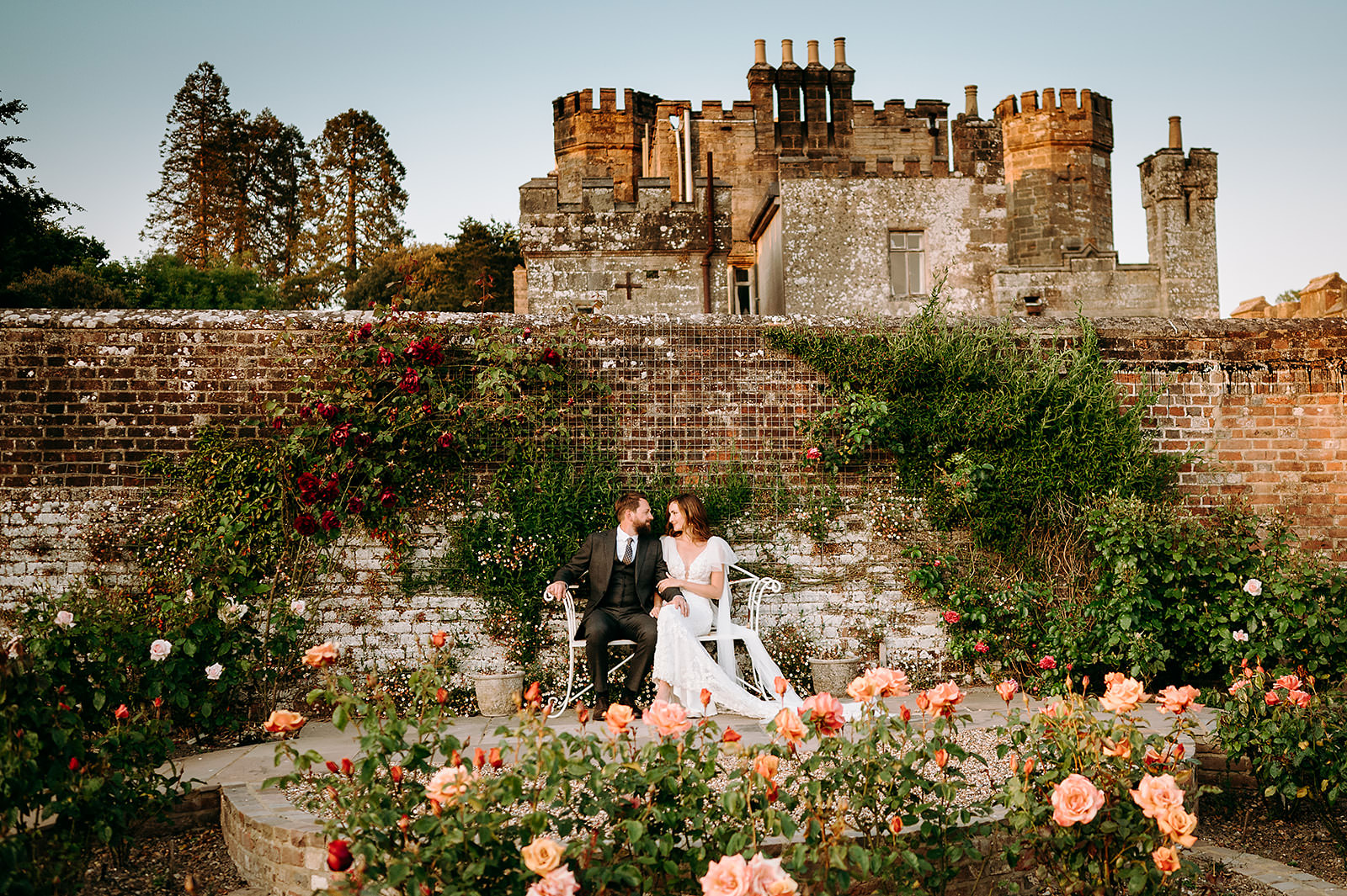 Bride and groom sitting on whiote bench surrounded by pink and ref flowerrs with castle in background